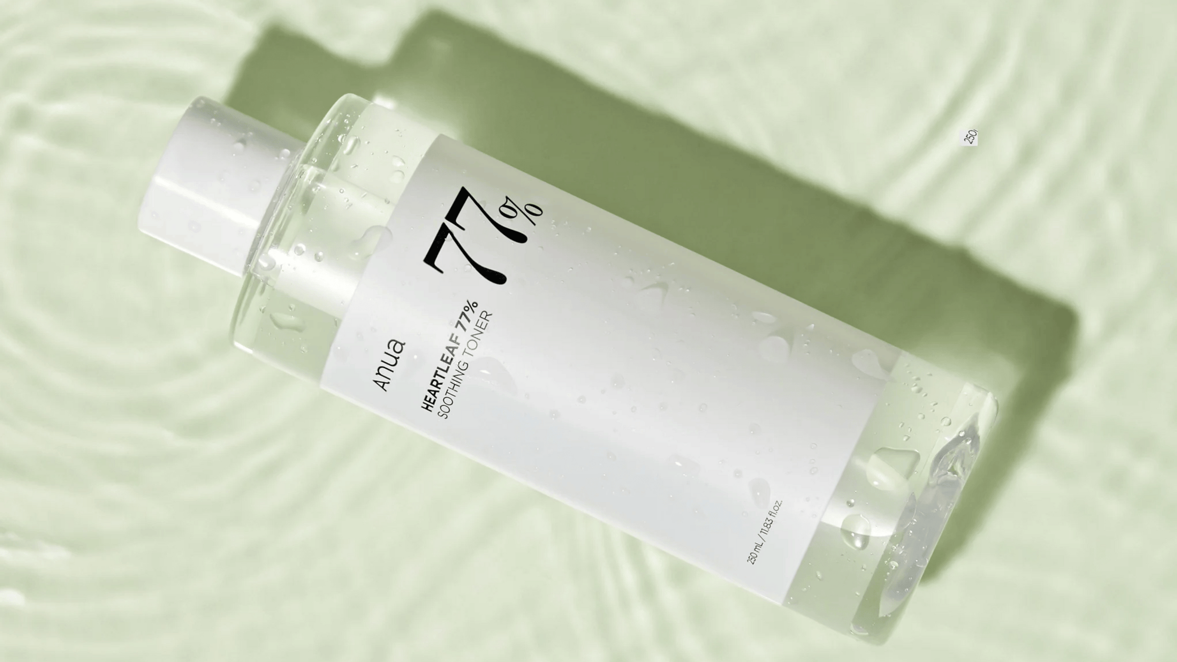 En genomskinlig flaska av Anua Heartleaf 77% Soothing Toner, täckt av vattendroppar, liggande på en texturerad ljusgrön bakgrund.