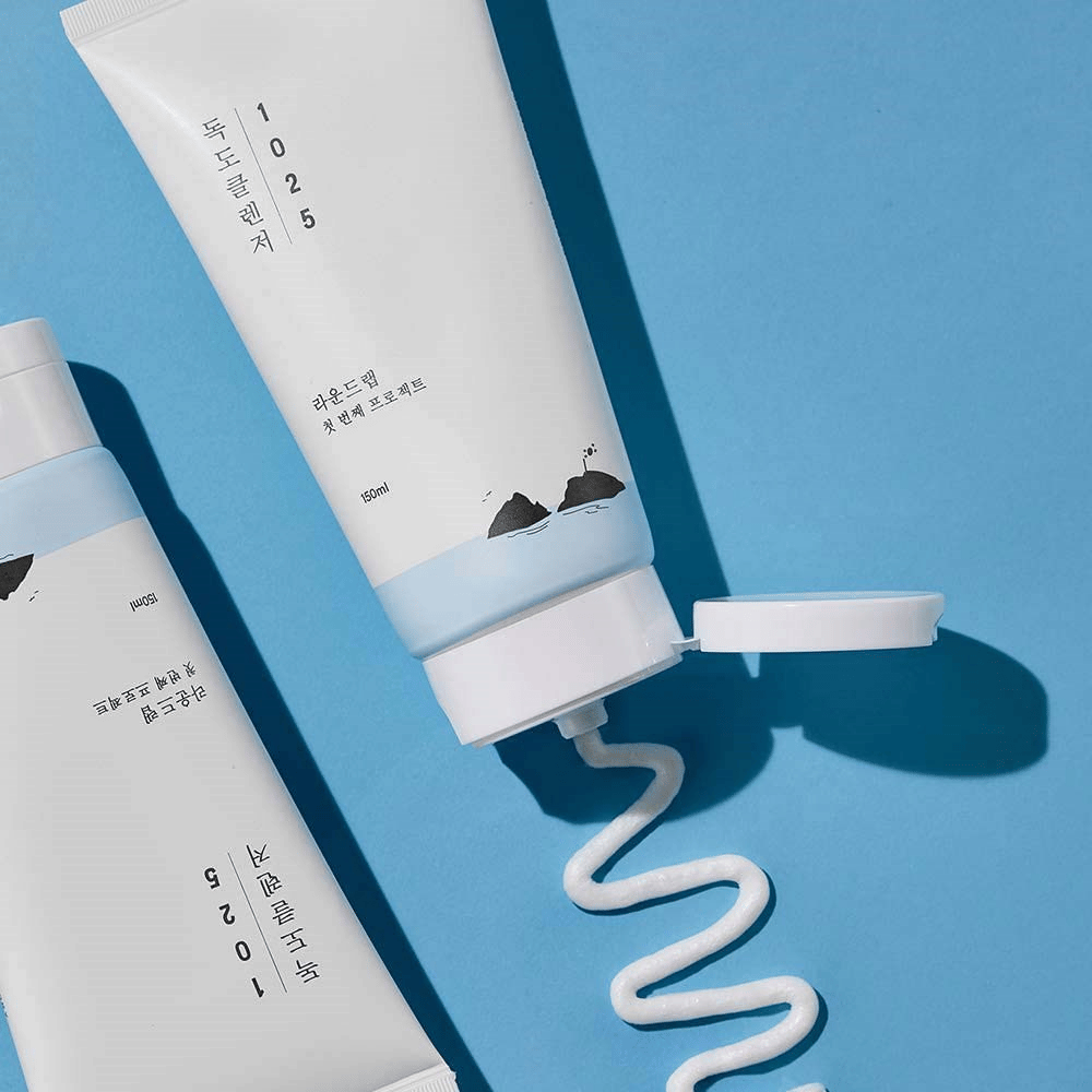 Kaksi tuubia 1025 Dokdo Cleanser sinisellä taustalla, joista yksi avoimella korkilla ja yksi aaltoileva puhdistusnauha puristettuna ulos.
