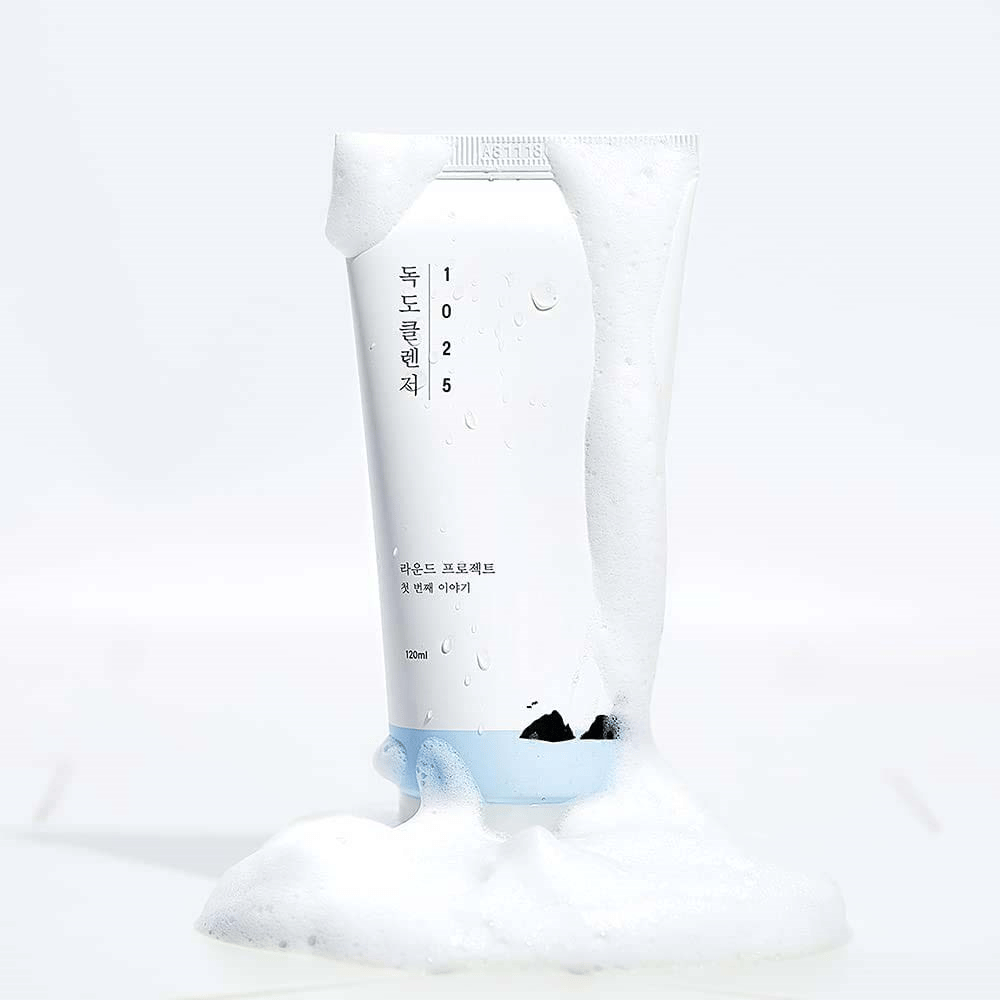 1025 Dokdo Cleanser valkoisessa tuubissa, joka on peitetty vaahdolla, vesipisaroilla ja vaahdolla pohjan ympärillä, irrotettu vaalealla taustalla.