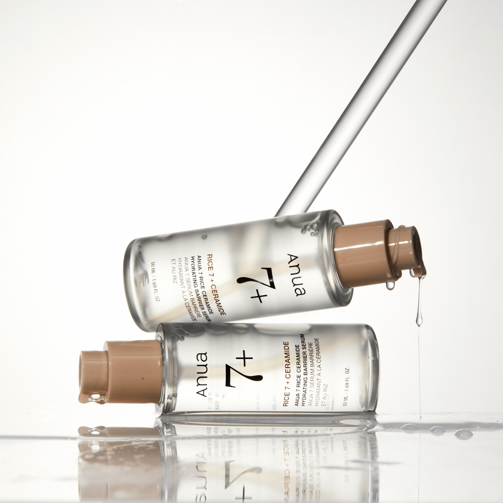 Kuvassa on kaksi läpinäkyvää pulloa "Anua 77 Rice + Ceramide Hydrating Barrier Serum" -seerumia, joista pisaroita valuu pipetistä. Riisillä ja keramideilla rikastettu seerumi on tarkoitettu kosteuttamaan ja vahvistamaan ihon suojakerrosta. Pullot on tyylikkäästi muotoiltu, mikä korostaa tuotteen premium-tunnetta.