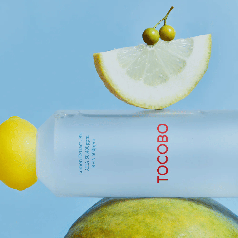TOCOBO AHA BHA Lemon Toner -pullo, koristeltu sitruunanmuotoisella korkilla. Sitruunaviipale ja pienet vihreät marjat tasapainottavat pullon päällä sinistä taustaa vasten.