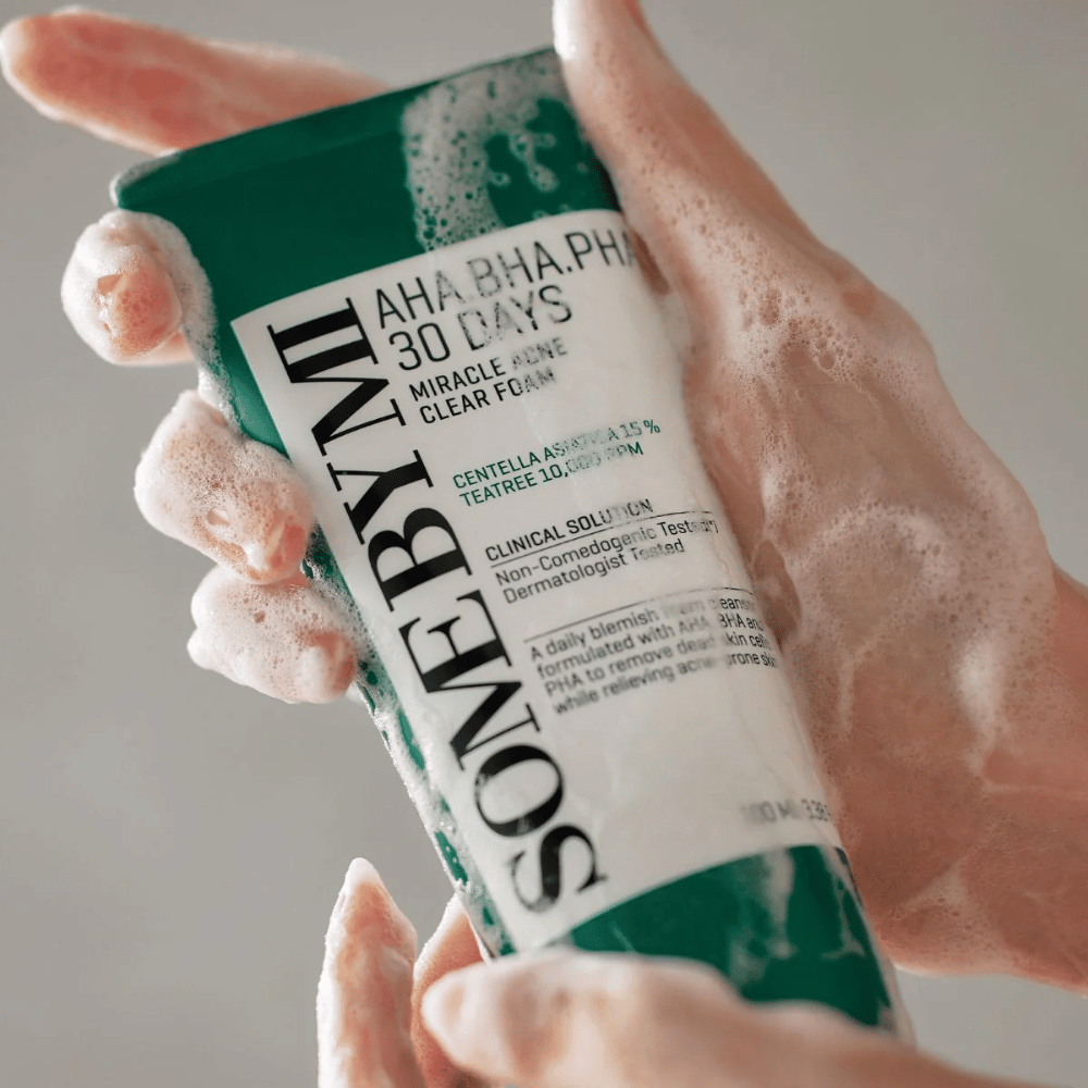 Henkilö pitää kädessään putkiloa "SOME BY MI AHA, BHA, PHA 30 Days Miracle Acne Clear Foam", jossa on vaahtoavaa sisältöä käsien päällä. Tuote on tarkoitettu päivittäiseen käyttöön aknen torjumiseksi ja huokosten puhdistamiseksi sen aktiivisilla ainesosilla, kuten centella, tea tree öljy ja hapot kuten AHA, BHA ja PHA.