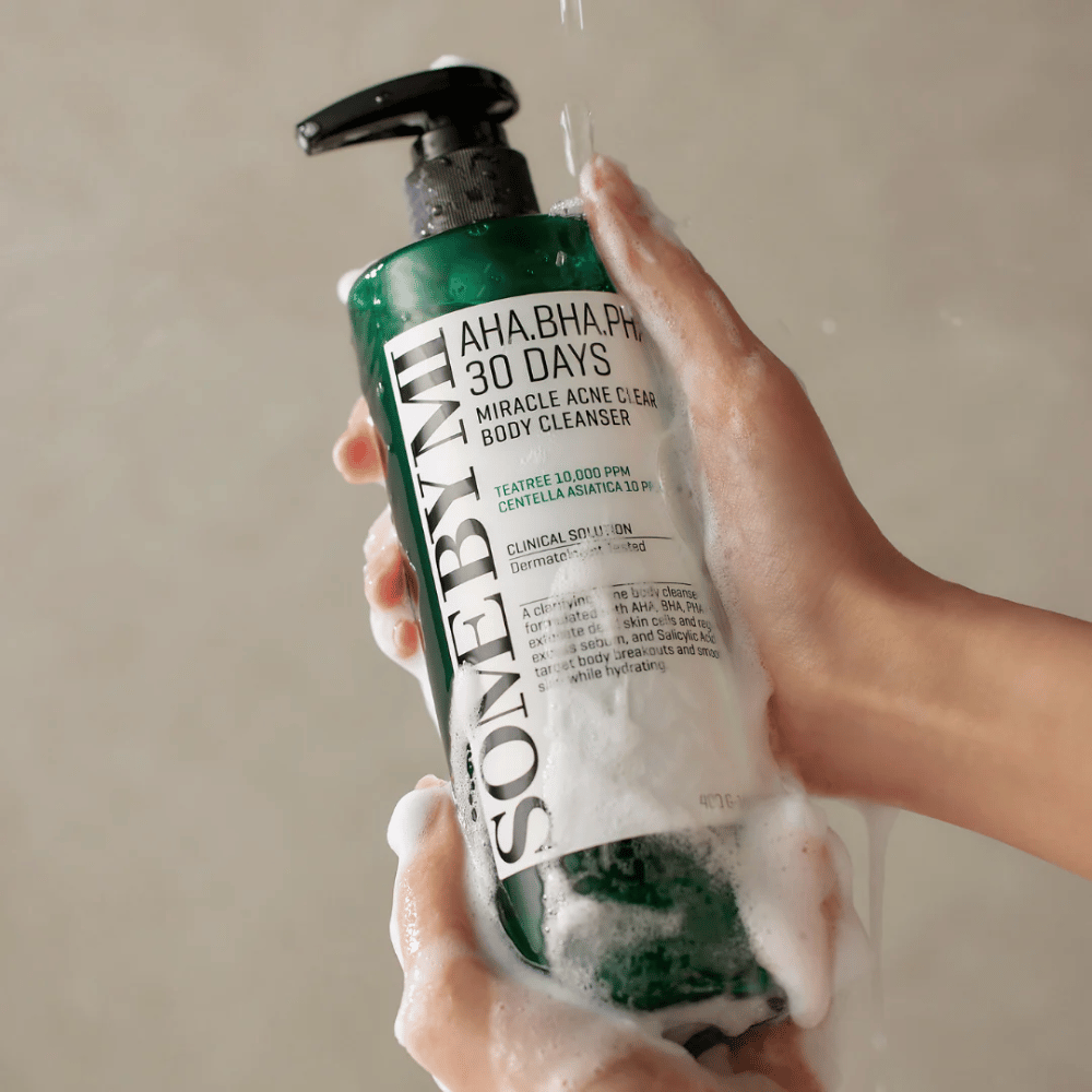 Henkilö pitää kädessään pulloa SOME BY MI AHA.BHA.PHA 30 Days Miracle Acne Clear Body Cleanser. Pullo on vihreä, valkoisella etiketillä ja se on peitetty puhdistusaineen vaahdolla. Henkilö pesee itseään tuotteella samalla kun vesi valuu käsille.