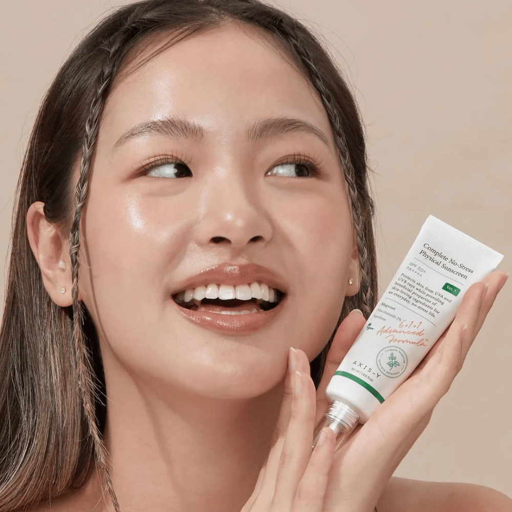Kuva esittää nuorta naista, joka hymyilee iloisesti ja pitelee tuubia, jossa on AXIS-Y Complete No-Stress Physical Sunscreen. Hänellä on säteilevä ja luonnollinen ulkonäkö vähäisellä meikillä, mikä korostaa esittelemäänsä tuotetta.