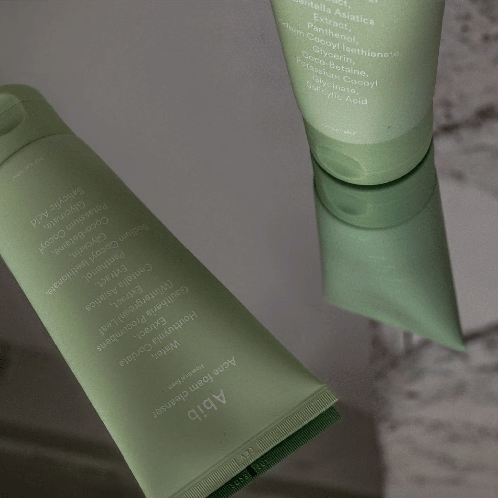 Vihreä tuubi Abib Acne Foam Cleanser Heartleaf makaa peilipöydällä tuotteen etupuoli ja teksti heijastettuna. Ainesosat kuten salisyylihappo, centella ja sydänlehtiuute näkyvät selvästi pakkauksessa.