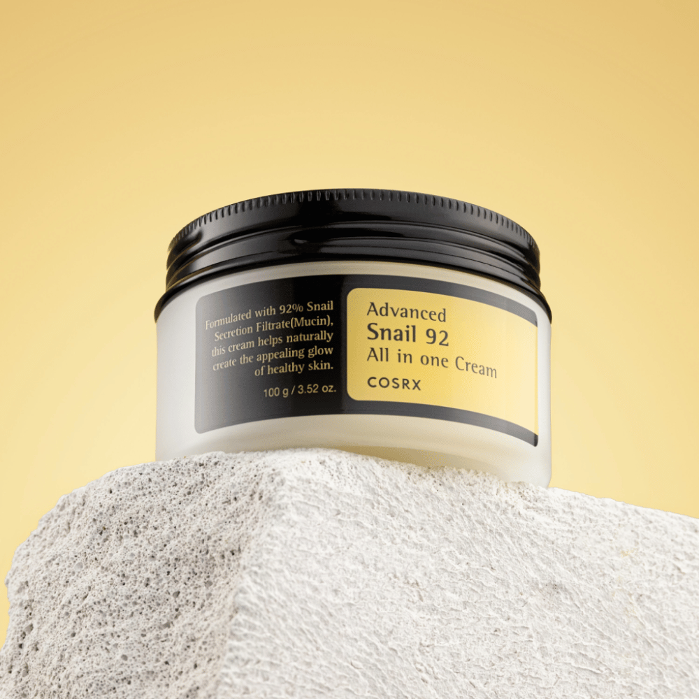 COSRX Advanced Snail 92 All in one Cream purkki sijoitettuna kiviselle pinnalle keltaisella taustalla.