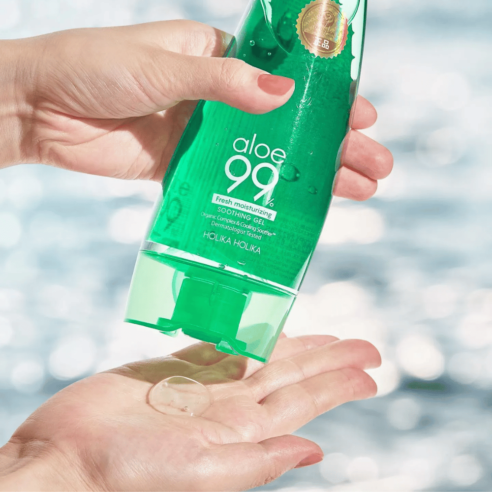 Käsi pitelee vihreää tuubia, jossa on Holika Holika Aloe 99% Soothing Gel, kun taas toinen käsi nappaa annosteltavan geelin. Tausta on sumea ja näyttää vetisen, kimaltelevan kuvion.