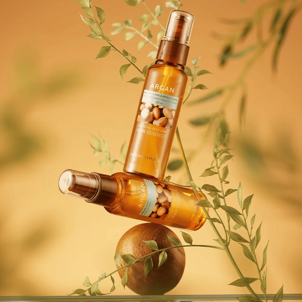 Kaksi pulloa Nature Republic Argan Essential Moisture Hair Oil asetettuna vihreiden lehtien joukkoon – silkinpehmeä kiilto kuiville hiuksille.