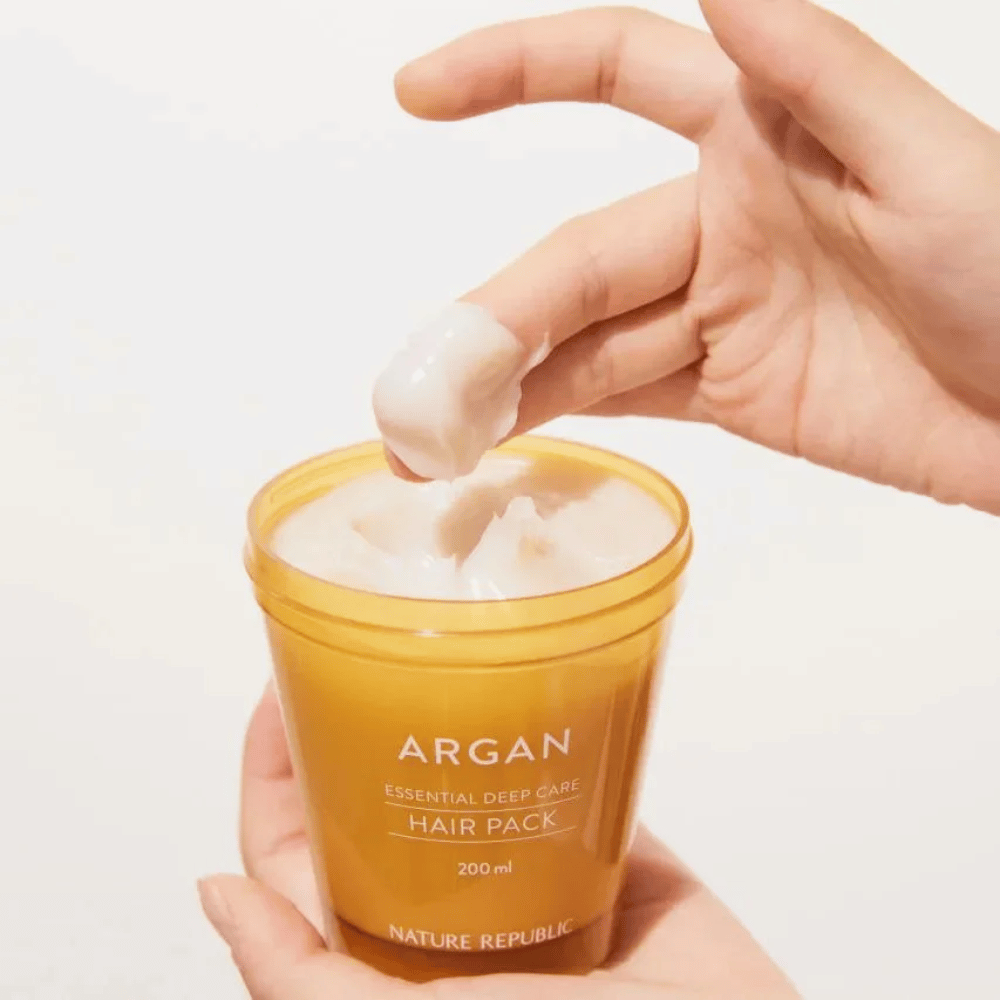 Käsi ottaa kermaista koostumusta Nature Republic Argan Essential Moisture Repairing Hair Mask – intensiivinen ravitseva hoito kuiville hiuksille.