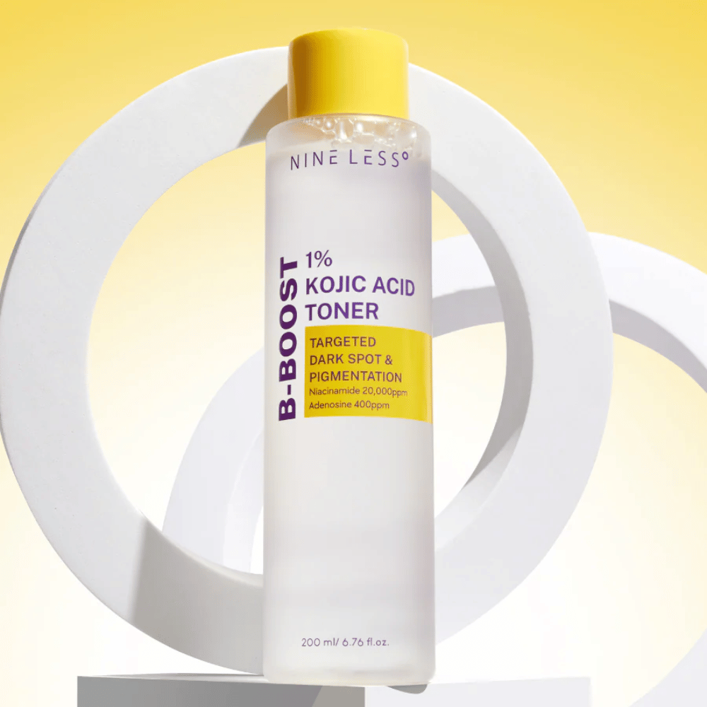 NINE LESS B-Boost 1% Kojic Acid Toner seisoo keltaista taustaa vasten tekstillä "Targeted Dark Spot & Pigmentation". Ihoa tasoittava toner, jossa on niasiiniamidia ja adenosiinia.