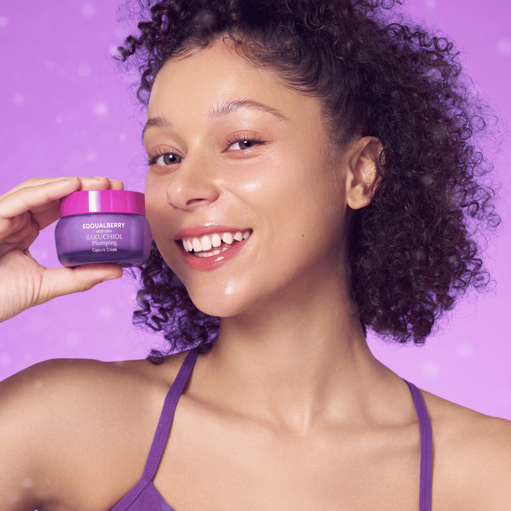 Nainen, jolla on hehkuva iho, pitää EQQUALBERRY Bakuchiol Plumping Capsule Cream ja hymyilee purppura taustaa vasten.