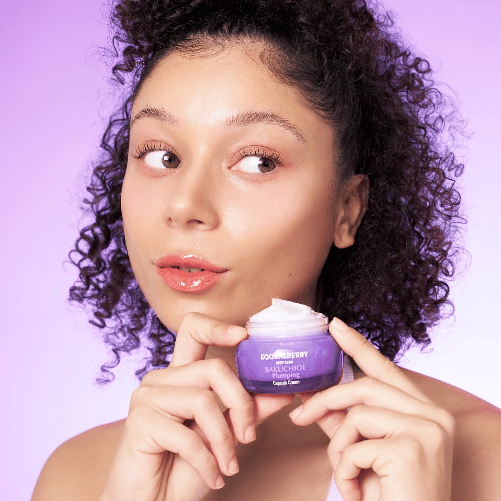 Nainen pitää EQQUALBERRY Bakuchiol Plumping Capsule Cream kädessään ja esittelee kosteutettua, hehkuvaa ihoaan purppura taustaa vasten.
