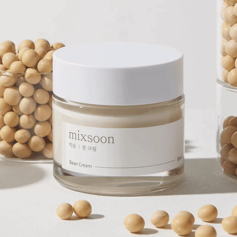 Läpinäkyvä purkki, jossa on valkoinen etiketti ja kansi, merkitty "mixsoon 팥 크림" ja "Bean Cream" tilavuudella "50ml". Purkki sisältää vaaleankeltaista voidetta ja on sijoitettu vaalealle pinnalle. Purkin vieressä on lasiastia täynnä vaaleanruskeita papuja, jotka ovat levinneet pinnalle, mikä viittaa siihen, että voide sisältää papu-uutetta. Tausta ja valaistus luovat lämpimän ja luonnollisen ilmapiirin.