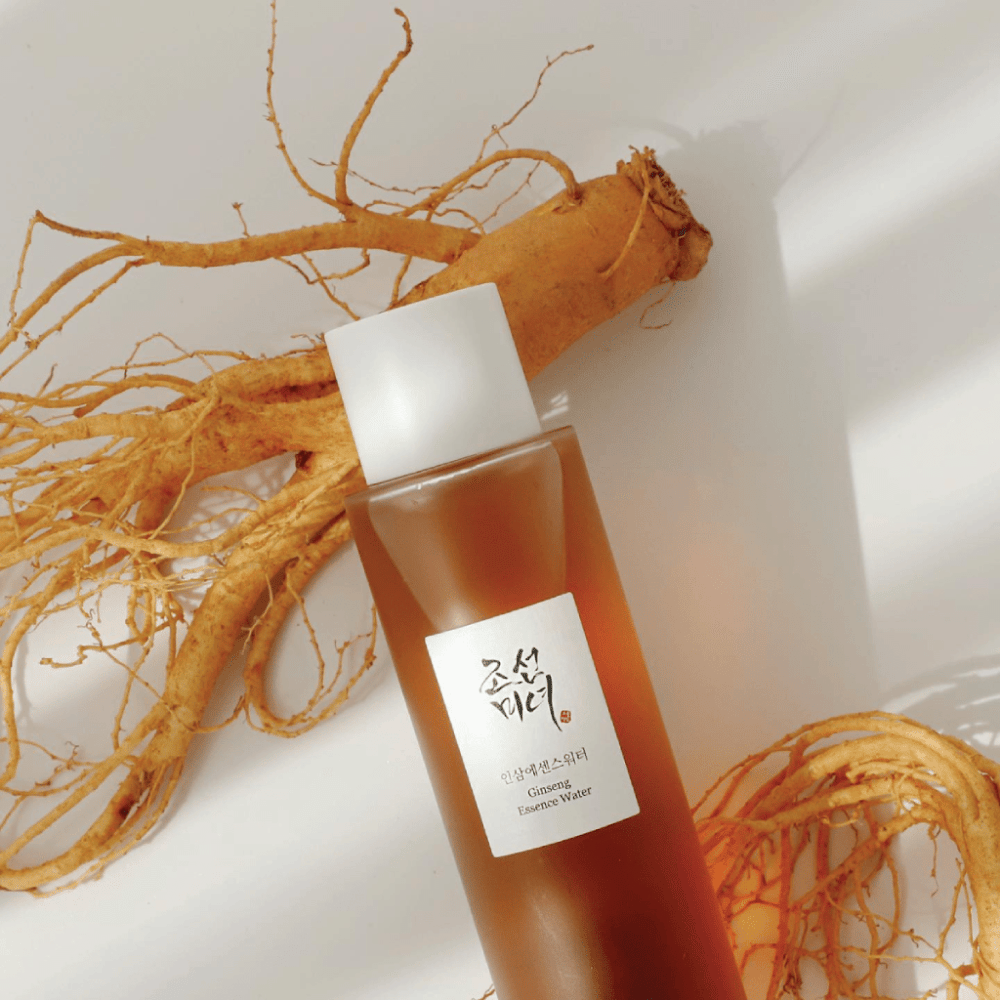 Ruskea pullo Beauty of Joseon Ginseng Essence Water valkoisella korkilla makaa tuoreen ginseng-juuren vieressä valkoisella taustalla.
