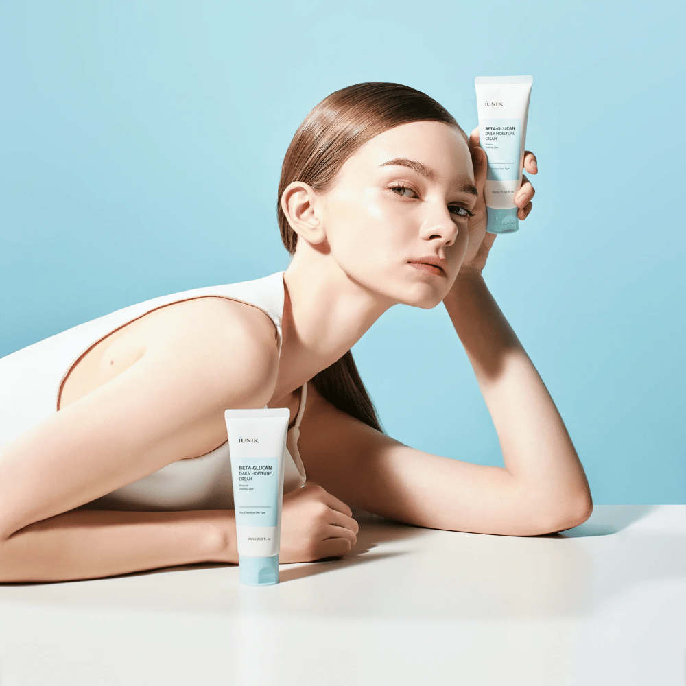 Lifestyle-kuva, jossa malli poseeraa tuotteen iUNIK Beta-Glucan Daily Moisture Cream kanssa vaaleansinistä taustaa vasten. Kuva välittää puhtaan, raikkaan ja kosteuttavan tunteen samalla, kun tuote on selkeästi esillä.