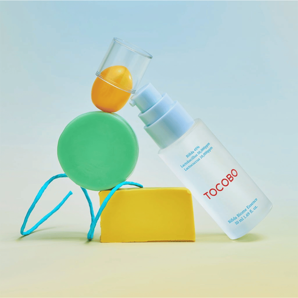TOCOBO Bifida Biome Essence huurretussa pumppupullossa, jossa on punainen logo, sijoitettuna värikkäälle taustalle, jossa on vihreitä, keltaisia ja oransseja geometrisia muotoja. Pullon teksti ilmoittaa Bifida 5 %, Lactobacillus 3 000 ppm ja Lactococcus 1 000 ppm.