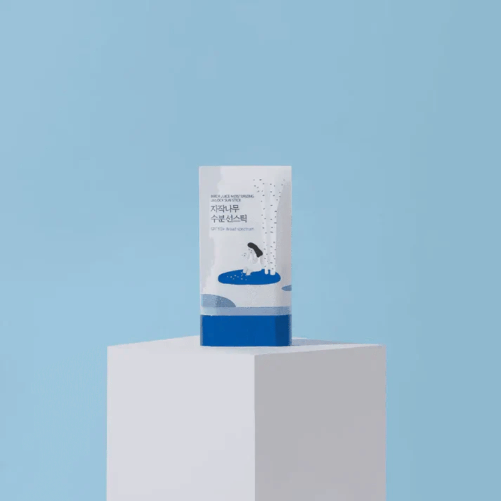 ROUND LAB Birch Juice Moisturizing Sun Stick SPF50+ seisoo jalustalla sinistä taustaa vasten – kosteuttava puikkomainen aurinkosuoja