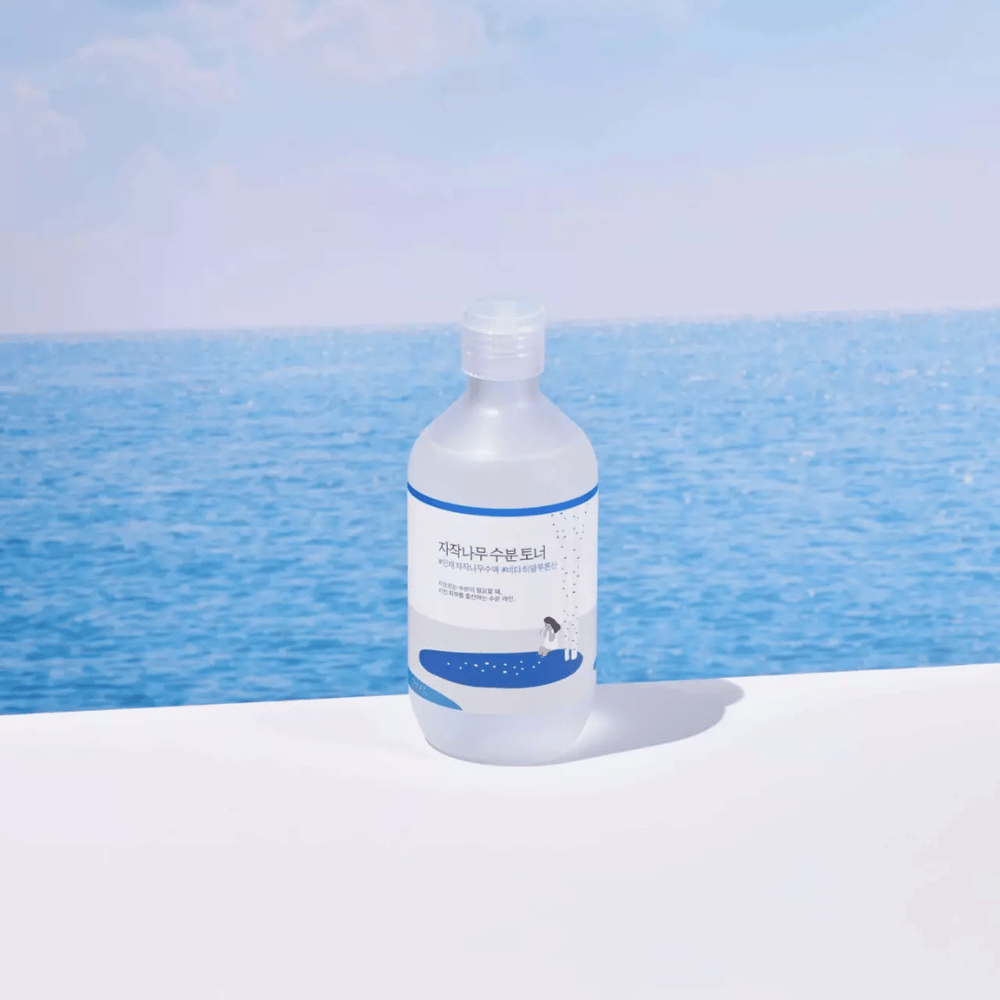 Pullo tuotetta ROUND LAB Birch Juice Moisturizing Toner seisoo valkoisella pinnalla, taustalla meri ja sininen taivas