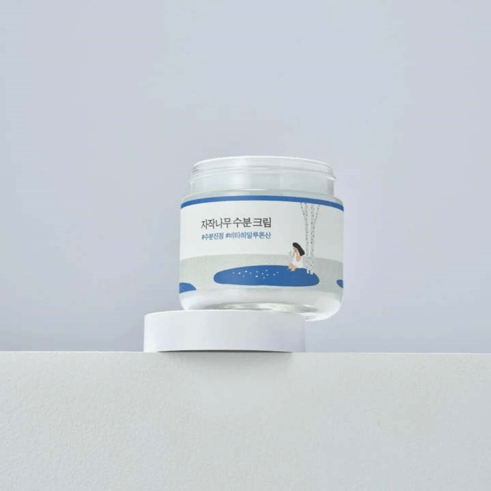 Round Lab Birch Juice Moisturizing Cream läpinäkyvässä purkissa, jossa on valkoinen etiketti ja sininen kuva naisesta koivun vieressä, seisoo valkoisella pinnalla vaaleaa taustaa vasten.
