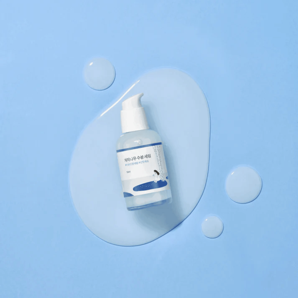 Round Lab Birch Juice Moisturizing Serum läpinäkyvässä pumppupullossa, sinisellä taustalla pisaroiden ja kaatuneen seerumin kanssa.
