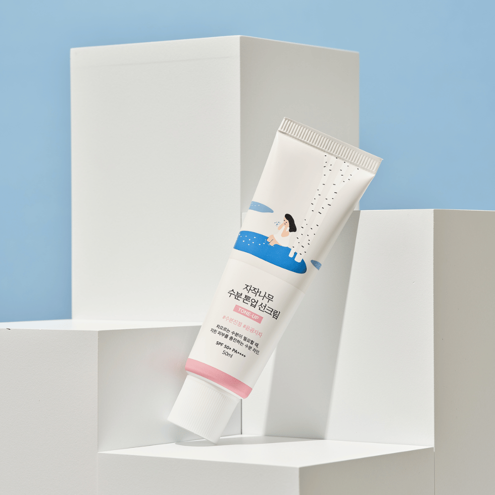 Putki, jossa on Birch Juice Moisturizing Tone Up Sunscreen, sijoitettuna valkoisille kuutioille vaaleansinisellä taustalla. Putki on valkoinen, ja siinä on kuva naisesta koivun ja veden äärellä. Koreankielinen teksti ilmoittaa, että tuote on SPF 50+ PA++++ ja sisältää 50 ml.
