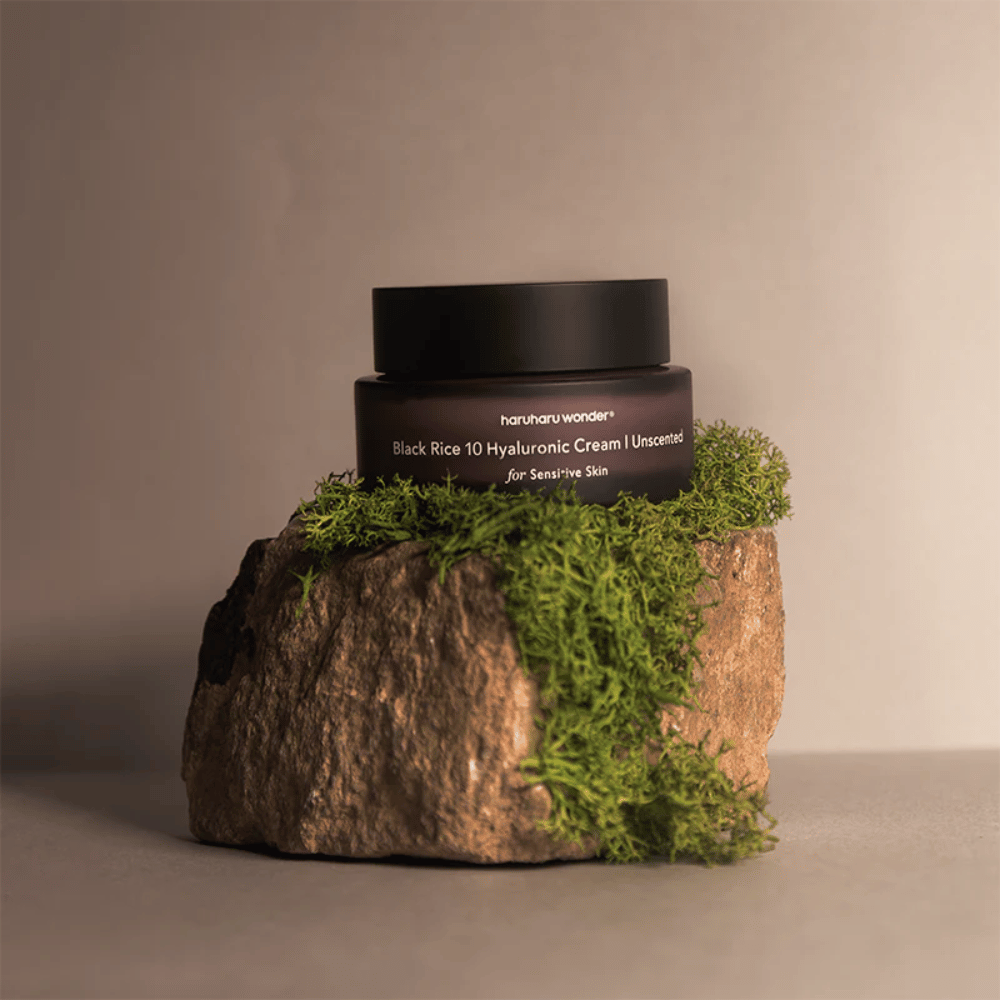 Kuva näyttää ihovoidepurkin, jossa on merkintä "haruharu wonder Black Rice 10 Hyaluronic Cream Unscented herkälle iholle". Purkki on musta ja seisoo suurella kivellä, joka on peitetty vaaleanvihreällä sammalella, mikä luo luonnollisen ja maanläheisen tunnelman.
