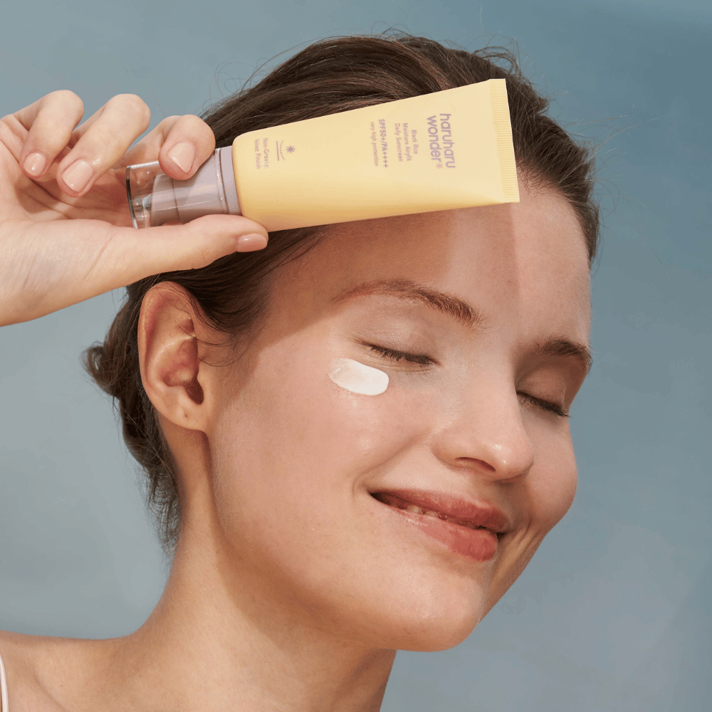Hymyilevä nainen levittää Black Rice Moisture Airyfit Daily Sunscreen SPF 50+ PA++++ kasvoilleen Haruharu Wonder:sta. Hän pitää kädessään keltaista aurinkovoideputkiloa ja hänellä on tuotetta poskellaan. Tausta on vaaleansininen.