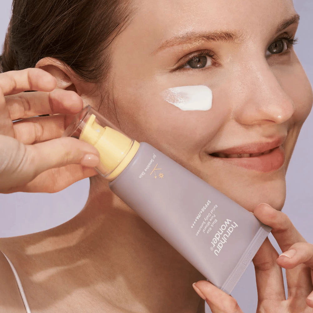 Hymyilevä nainen levittää Haruharu Wonder Black Rice Pure Mineral Relief Daily Sunscreen SPF 50+ PA++++ kasvoilleen. Hän pitää käsissään lila aurinkovoideputkiloa ja hänellä on tuotetta poskellaan. Tausta on vaaleanlila.