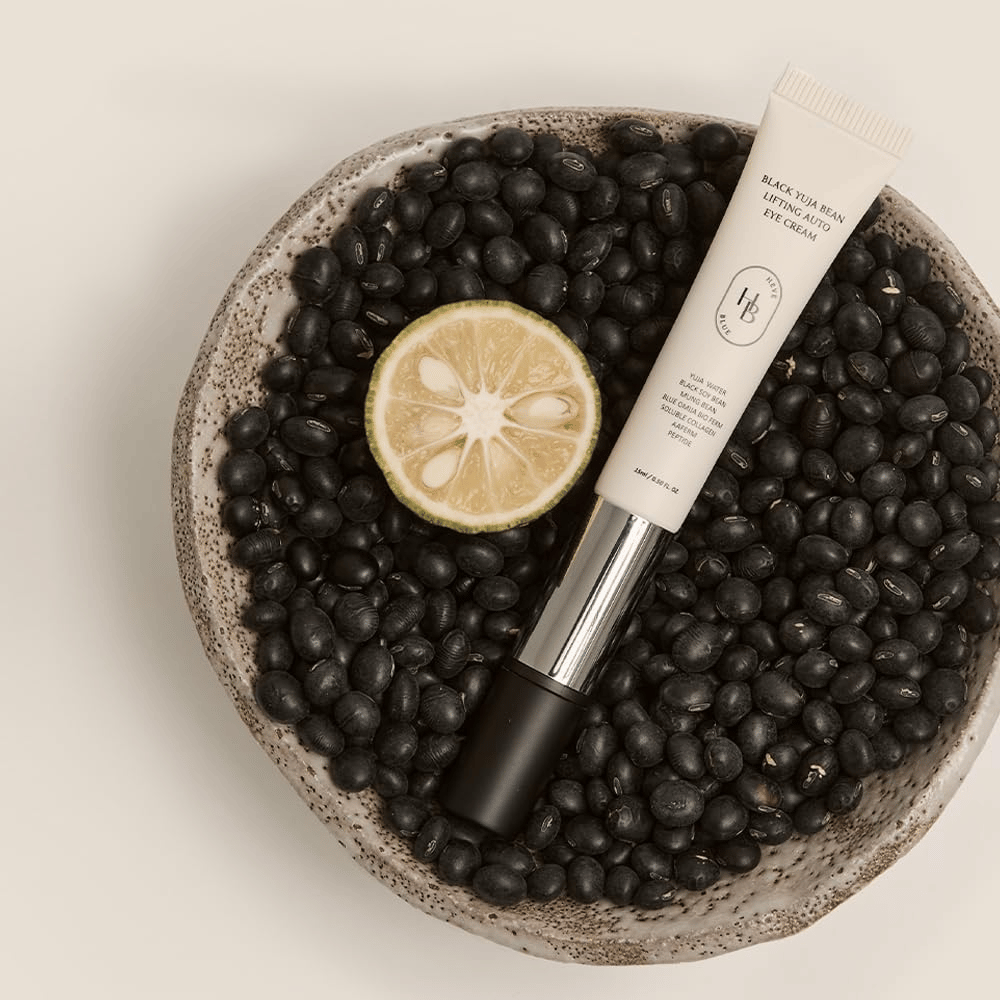 Black Yuja Bean Lifting Auto Eye Cream makaa kulhossa, joka on täynnä mustia papuja ja puolikas sitrushedelmä – visuaalinen esitys tuotteen luonnollisista ainesosista.