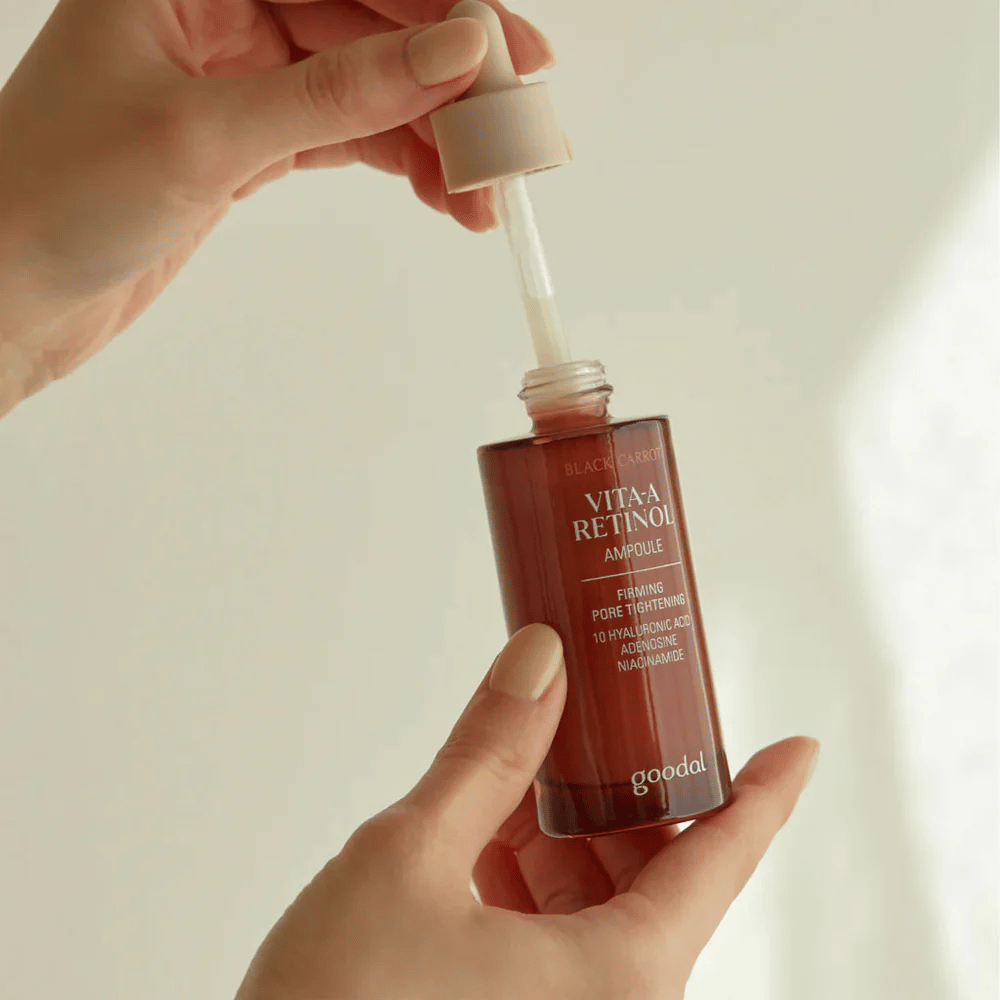 Käsi pitää pulloa, jossa on Goodal Black Carrot Vita-A Retinol Firming Ampoule. Pullon etiketti näyttää tuotteen nimen ja ominaisuudet, mukaan lukien "Firming Pore Tightening" ja ainesosat kuten 10% hyaluronihappo, adenosiini ja niasiiniamidi. Pipetti on väriltään beige.