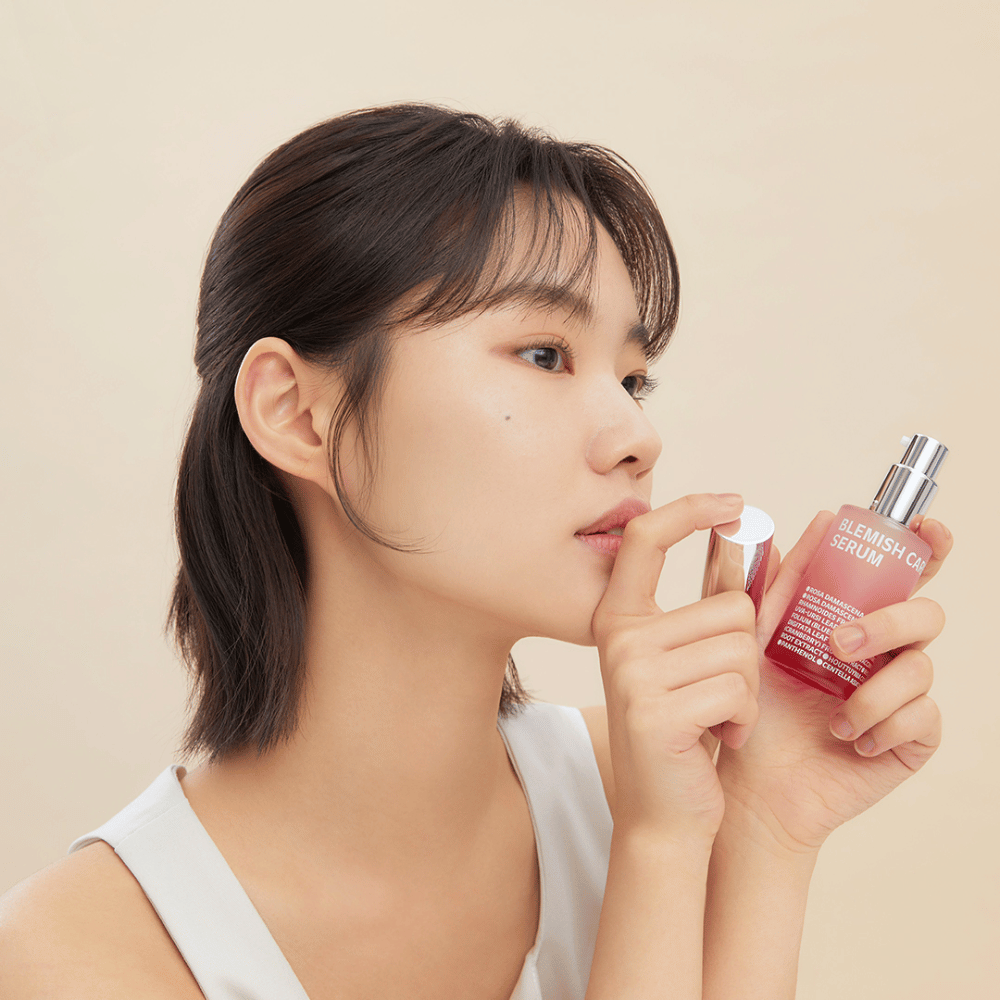 Nainen, jolla on tummat hiukset, pitää Blemish Care Serum -pulloa yhdessä kädessä ja irrottaa korkin toisella kädellä. Hänellä on neutraali ilme ja tausta on vaalea beige, mikä antaa pehmeän ja minimalistisen vaikutelman.