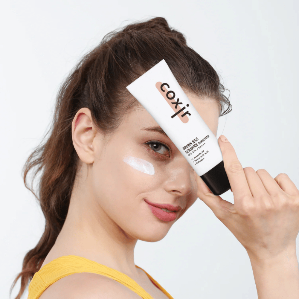 Nainen, jolla on aurinkovoidetta poskellaan, pitää ylhäällä tuubia Coxir Brown Rice Ceramide Sunscreen, SPF 50+ PA+++, otsaansa vasten ja hymyilee kevyesti.