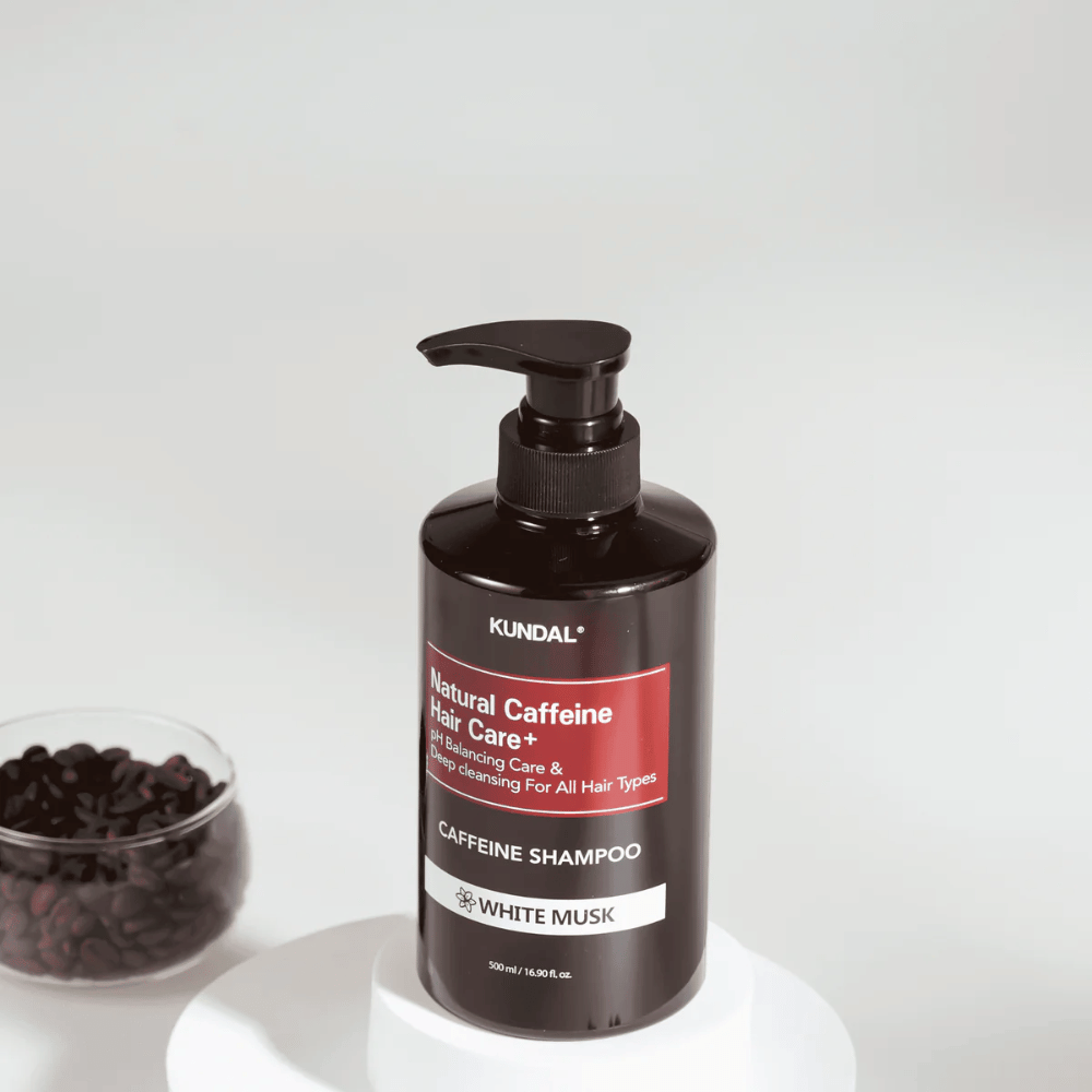 KUNDAL Natural Caffeine Hair Care+ Shampoo White Musk mustassa pumppupullossa, 500 ml, sijoitettu kahvipapukulhon viereen.
