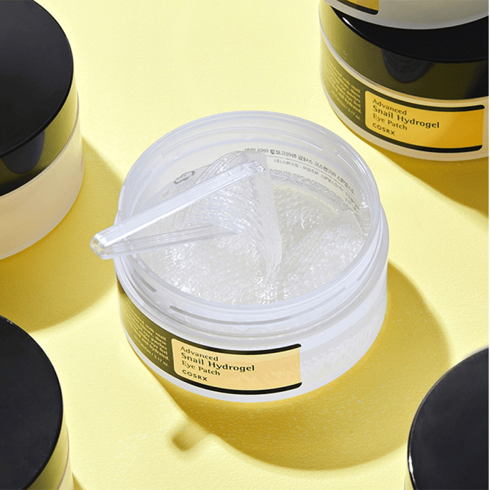 Avoin purkki COSRX Advanced Snail Hydrogel Eye Patch läpinäkyvillä silmälaastareilla ja pienellä lastalla, ympäröitynä useilla suljetuilla purkeilla keltaisella taustalla.