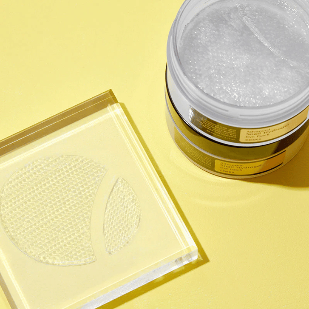 Avoin purkki COSRX Advanced Snail Hydrogel Eye Patch seisoo keltaisella taustalla läpinäkyvän alustan vieressä, jossa on kaksi silmälaastaria.
