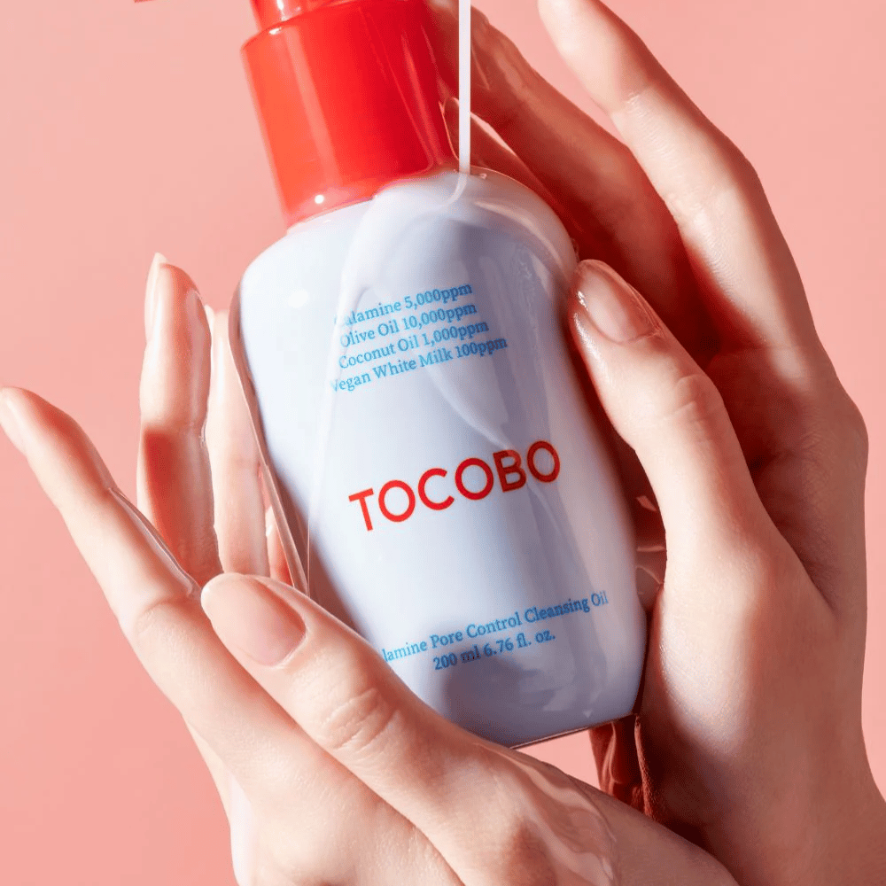 TOCOBO Calamine Pore Control Cleansing Oil pidetään käsissä vaaleanpunaista taustaa vasten, pullon punainen pumppu ja ainesosaluettelo näkyvät selvästi.