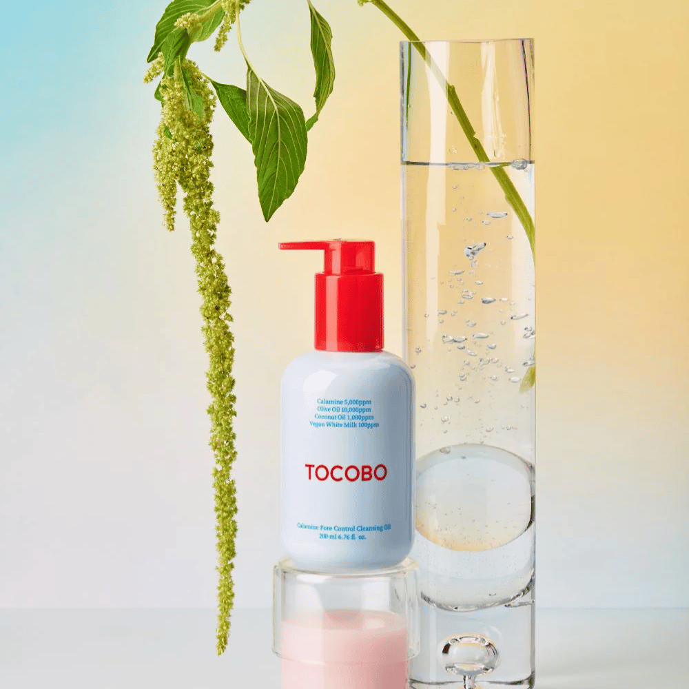 TOCOBO Calamine Pore Control Cleansing Oil punaisella pumpulla seisoo korkean vesitäytetyn lasiputken ja vihreän kasvin vieressä, vaalean pastellinväristä taustaa vasten.