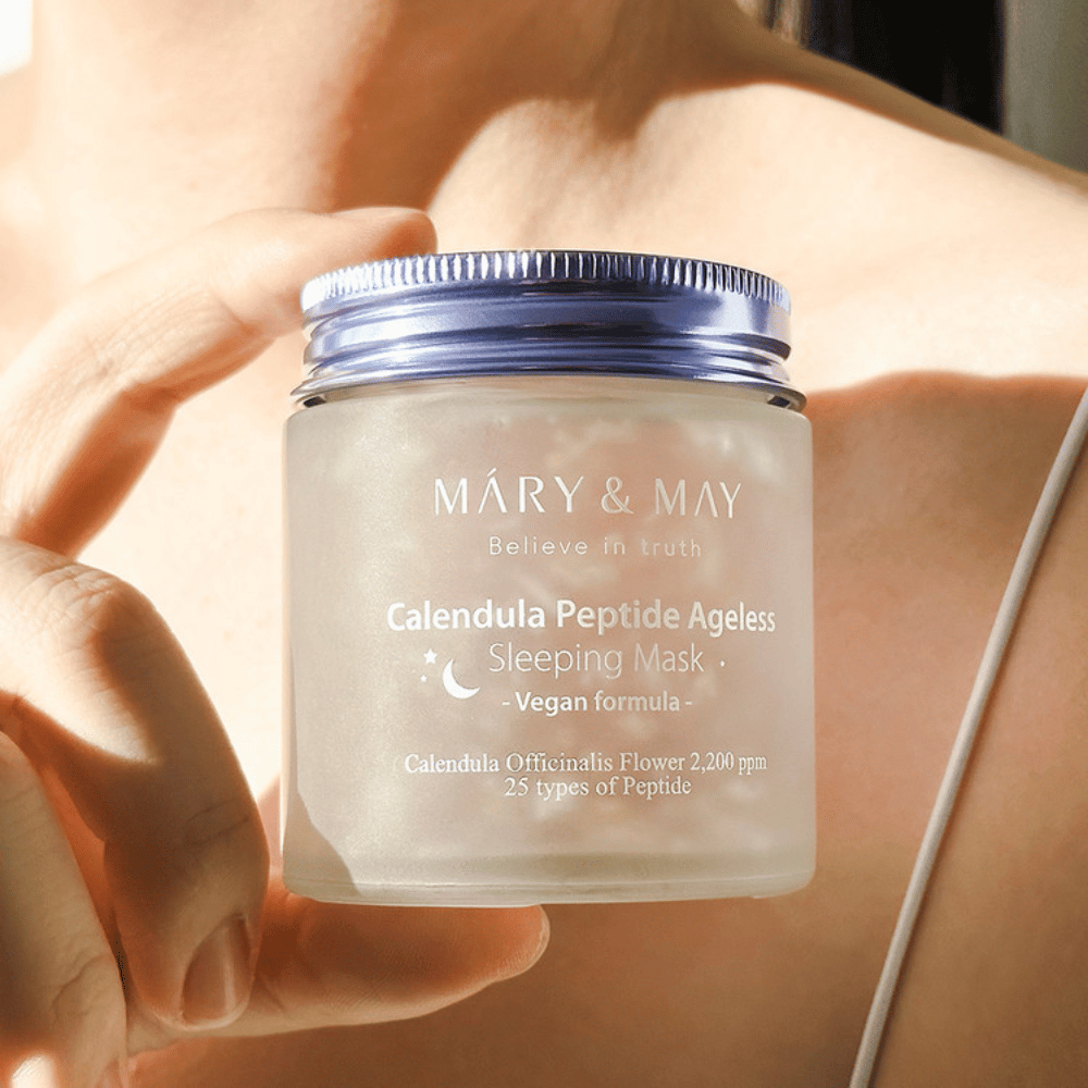 Nainen pitää kädessään purkkia, jossa on MARY & MAY Calendula Peptide Ageless Sleeping Mask. Purkki on läpinäkyvä ja siinä on violetti kansi, ja teksti "Calendula Peptide Ageless Sleeping Mask" näkyy selvästi. Tuote on vegaaninen yönaamio, joka sisältää kehäkukkaa ja peptidejä antaakseen iholle nuorekasta hehkua yön aikana.