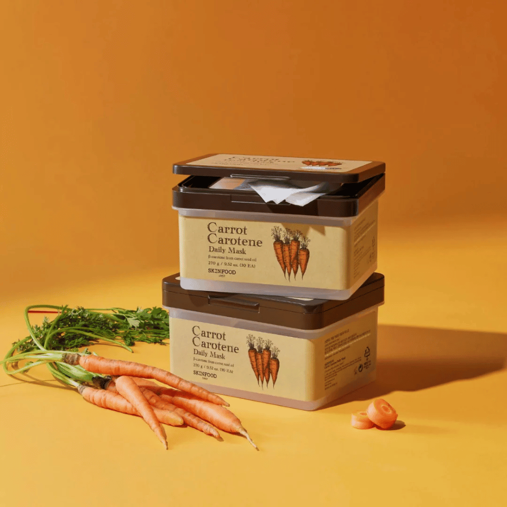 Kuvassa on kolme pinottua "Carrot Carotene Daily Mask"-pakkausta SKINFOOD:stä, asetettuna oranssille taustalle tuoreiden porkkanoiden kanssa. Jokainen laatikko sisältää kasvonaamioita, jotka on rikastettu porkkanansiemenöljystä saadulla β-karoteenilla, suunniteltu päivittäiseen käyttöön ihon ravitsemiseksi ja hoitamiseksi.