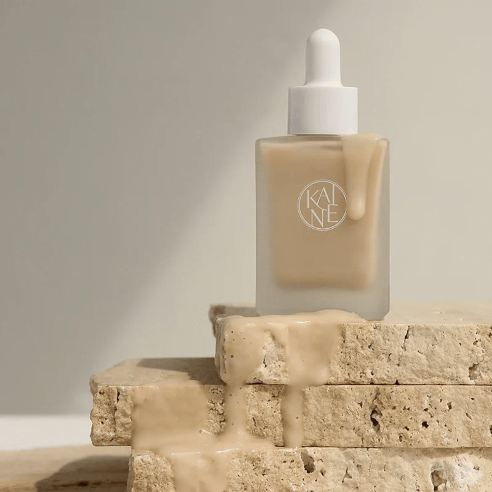 KANE Chaga Collagen Charging Serum huurretussa lasipullossa, jossa on pipetti. Kermainen koostumus valuu luonnollisten kivien yli pehmeässä ja minimalistisessa valossa.