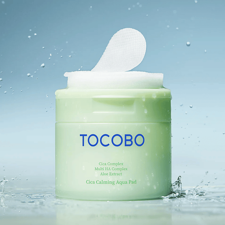 Avattu purkki tuotetta TOCOBO Cica Calming Aqua Pad, jossa yksi toner pad -lappu kohoaa pakkauksesta. Vesipisarat tuotteen ympärillä vahvistavat intensiivisen kosteutuksen ja raikkauden tunnetta.