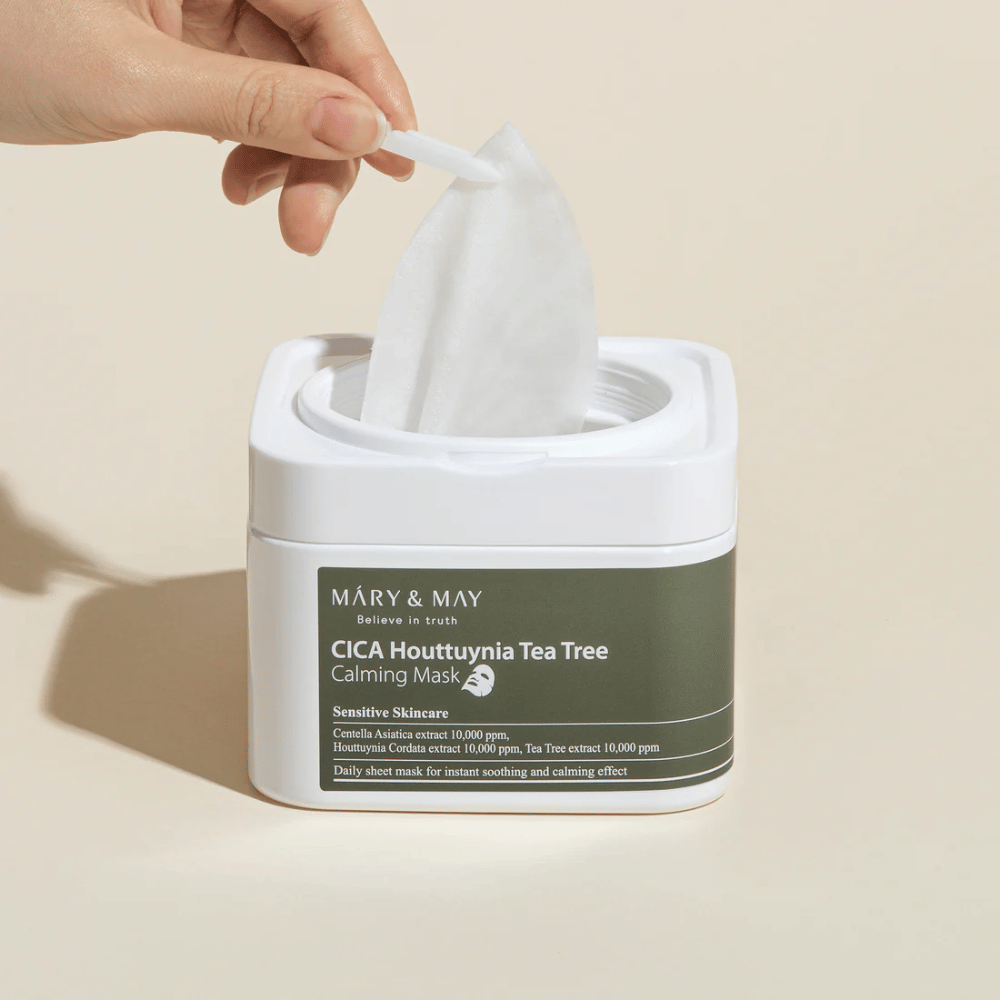 Käsi nostaa sheetmaskin Mary & May Cica Houttuynia Tea Tree Calming Mask-purkista – rauhoittava kasvonaamio herkälle iholle Cican ja Tea Tree-uutteen kanssa.