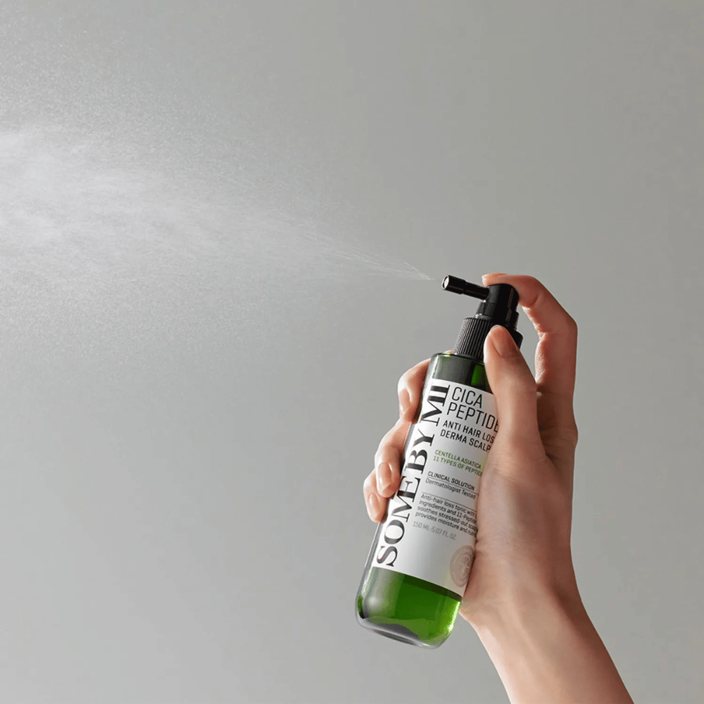 Käsi suihkuttaa SOME BY MI Cica Peptide Anti Hair Loss Derma Scalp Tonic vihreästä pullosta mustalla suuttimella ja valkoisella etiketillä harmaata taustaa vasten.