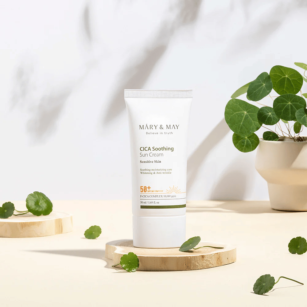 Mary&May CICA Soothing Sun Cream asetettuna puukorokkeelle, jota ympäröivät vihreät centella-lehdet. Kuva korostaa ihoa rauhoittavia ainesosia ja tuotteen taustalla olevaa luonnollista inspiraatiota.