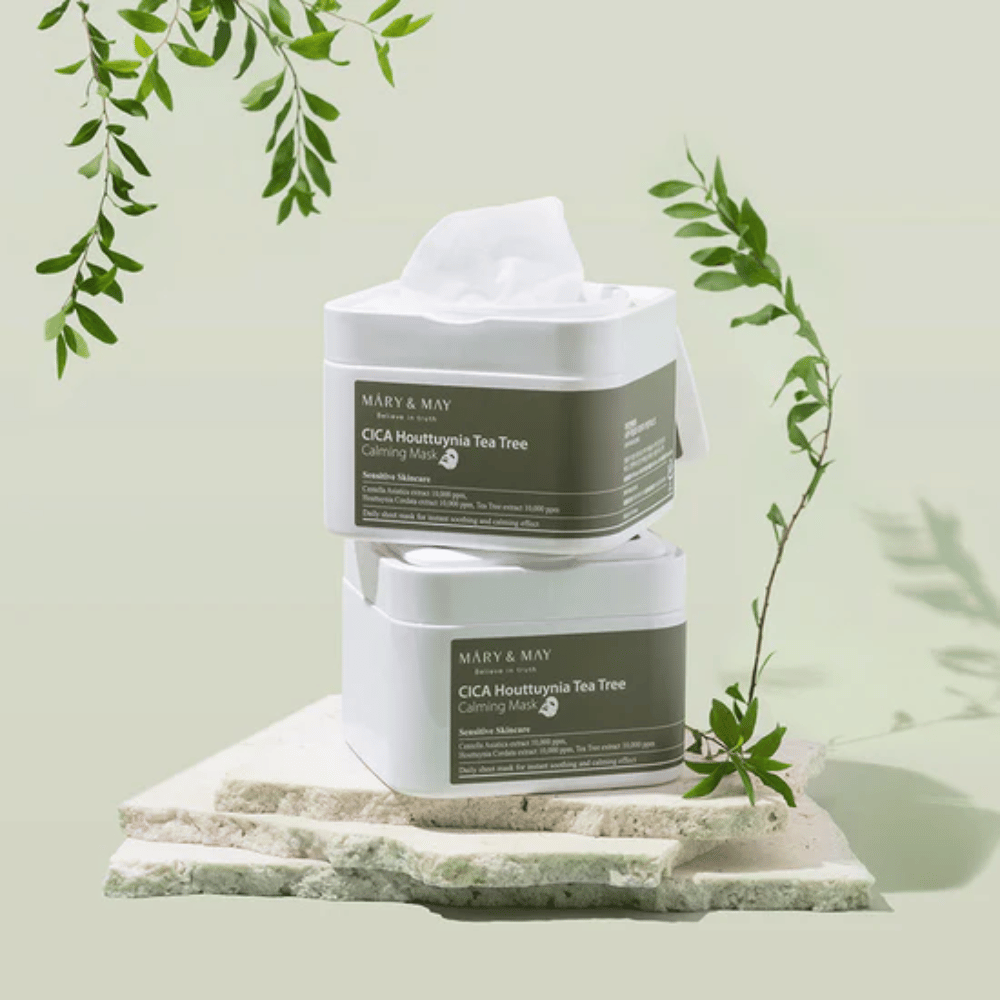 Kaksi Mary & May Cica Houttuynia Tea Tree Calming Mask-purkkia asetettuna luonnonkivelle vihreillä kasvidetaljeilla – rauhoittavia sheetmaskeja herkälle iholle.