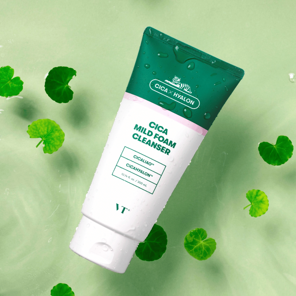 VT Cica Mild Foam Cleanser makaa vaaleanvihreällä alustalla, jossa on vesipisaroita pakkauksessa ja jota ympäröivät vihreät centella asiatica -lehdet, mikä korostaa tuotteen rauhoittavia ominaisuuksia.