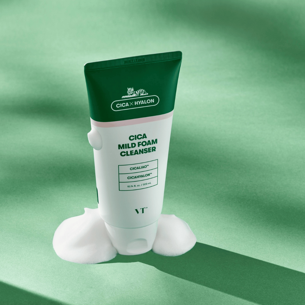 VT Cica Mild Foam Cleanser -tuubi seisoo vihreällä alustalla, jossa on vaahtoa pohjan ympärillä. Tuotteessa on vihreä ja valkoinen muotoilu ja sitä markkinoidaan CICALIAO™ ja CICAHYALON™ -aineilla.
