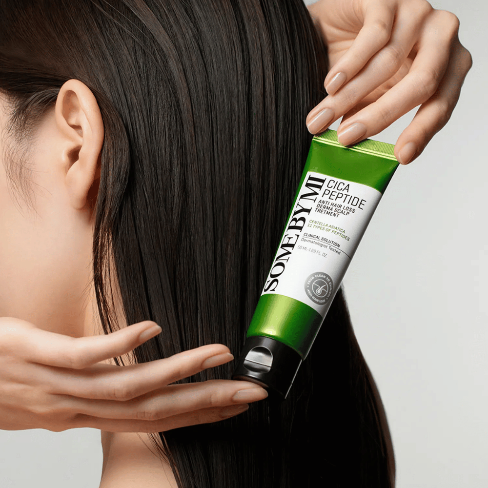Nainen levittää SOME BY MI Cica Peptide Anti Hair Loss Derma Scalp Treatment pitkään, tummaan hiukseen vihreällä ja valkoisella tuubilla.