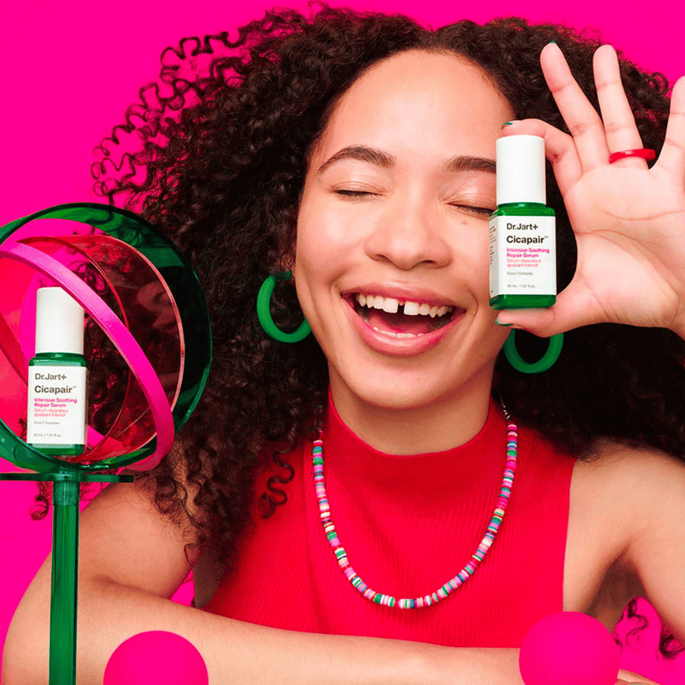 Nainen hymyilee leveästi pitäessään Dr.Jart+ Cicapair Intensive Soothing Repair Serum kasvojensa edessä. Hänellä on vihreät korvakorut ja punainen kaulakoru. Tausta on vaaleanpunainen ja koristeltu vihreillä ja vaaleanpunaisilla ympyröillä.