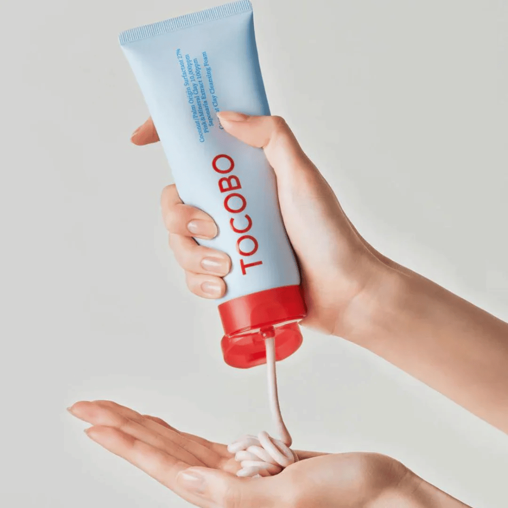 Henkilö pitää kädessään TOCOBO Coconut Clay Cleansing Foam -tubin. Tubi on vaaleansininen, jossa on punainen korkki ja teksti "TOCOBO" edessä. Tuotetta puristetaan ulos tubista ja se muodostuu shampooksi kädessä. Tausta on neutraali ja korostaa tuotetta.